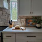 Austin Rustic Ashlar natural thin stone veneer installed as a kitchen backsplash. The blend's multi-tone cream, tan, and grey-buff stones surround a window above the range, paired with white shaker cabinets, brass hardware, and a light quartz counter. Real customer installation — sold by Stoneyard, Inc., Littleton, MA.