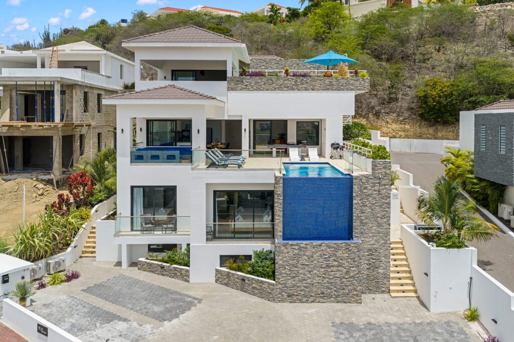 Drone view of a white modern villa in Curaçao with a stacked stone wall beneath a blue tiled balcony pool.