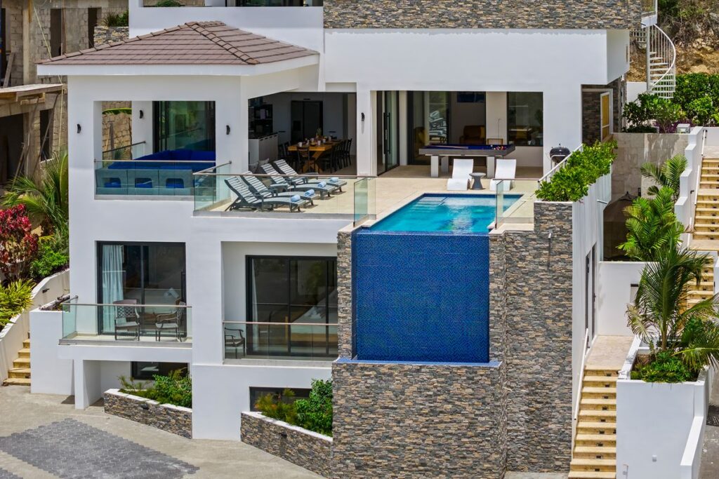 Front view of a white villa with a blue tiled pool on a wall clad in multicolored stacked stone panels.