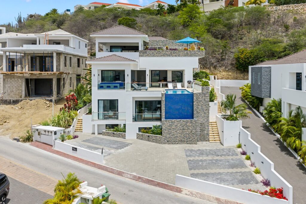Hillside villa in Curaçao with stacked stone walls, a blue balcony pool and stairs beside a paved driveway.