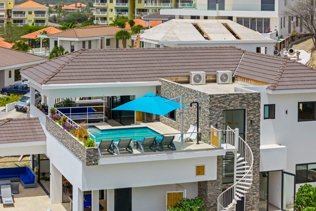 Aerial view of a rooftop pool with a turquoise umbrella, stacked stone parapet walls and a white spiral staircase.