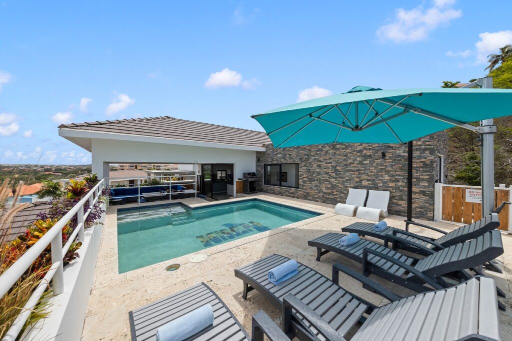 Pool deck with loungers and a turquoise umbrella in front of a pavilion with a stacked stone side wall.