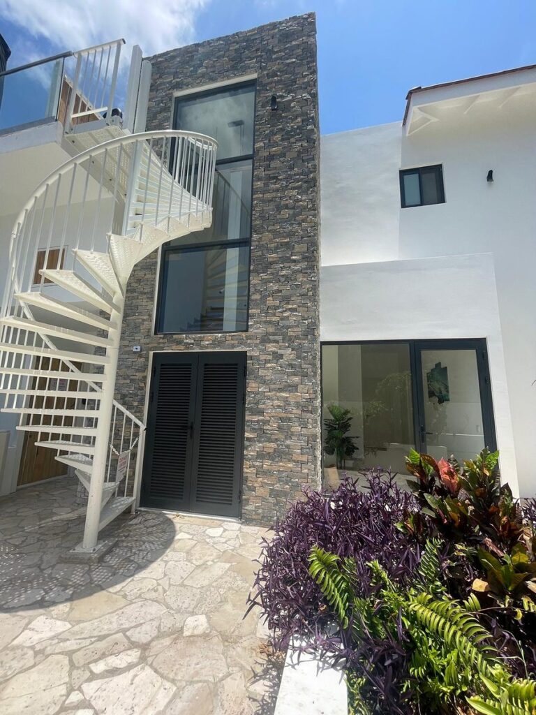 Tall stacked stone tower with glass windows next to a white spiral staircase and tropical plants.