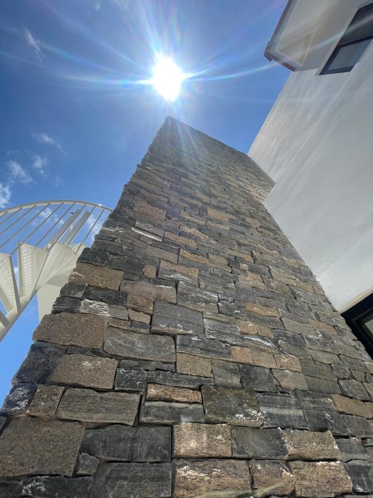 Upward view of a stacked stone column against a bright blue sky with the sun above.