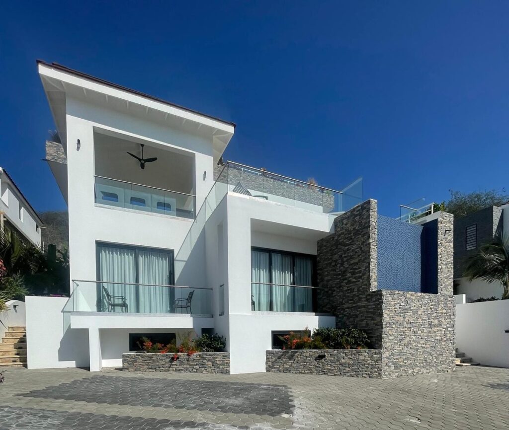 Street view of a white modern villa with stacked stone walls and planters under a deep blue sky.
