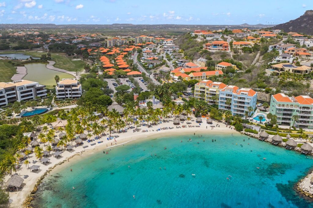 Aerial view of a curved white sand beach with turquoise water, palm trees and orange-roofed resort buildings.